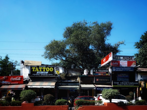 A street scene with multiple shops and signage, including a tattoo parlor offering discounts, a Coca-Cola advertisement, and various other small businesses. The area is lined with trees and shrubs, and there are parked vehicles in front.