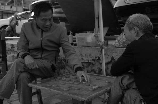 Two elderly men are engaged in playing a game of Chinese checkers or a similar strategy game on a street-side setup. One man is making a move while the other observes. The scene takes place outdoors, with vehicles and various items like a bag with heart patterns visible in the background, creating a casual, everyday urban atmosphere.