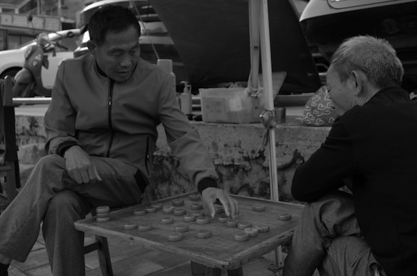 Two elderly men are engaged in playing a game of Chinese checkers or a similar strategy game on a street-side setup. One man is making a move while the other observes. The scene takes place outdoors, with vehicles and various items like a bag with heart patterns visible in the background, creating a casual, everyday urban atmosphere.