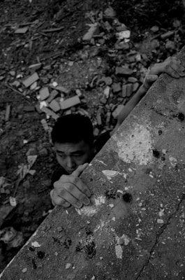 A black and white image of a person gripping the edge of a rough, somewhat damaged concrete ledge. The ground below is strewn with debris, bricks, and rubble.