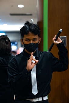 A person is dressed in a black outfit with a white tie and a white belt, wearing a black mask and holding a sword over their shoulder. The background shows an indoor environment with a green vertical stripe on the wall.