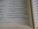 A close-up view of an open book showing text laid out in verses or poetic form. The text appears to be printed in black ink on off-white pages.