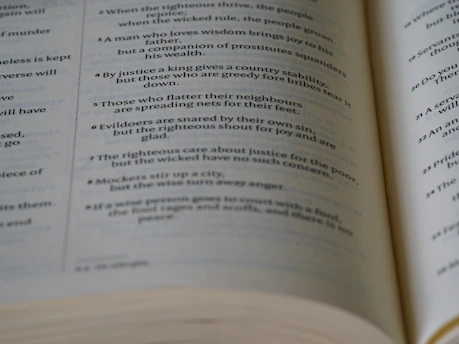 A close-up view of an open book showing text laid out in verses or poetic form. The text appears to be printed in black ink on off-white pages.