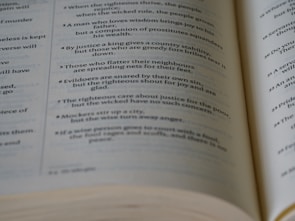 A close-up view of an open book showing text laid out in verses or poetic form. The text appears to be printed in black ink on off-white pages.