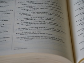 A close-up view of an open book showing text laid out in verses or poetic form. The text appears to be printed in black ink on off-white pages.