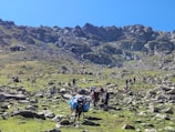 A rugged mountain landscape with rocky terrain and patches of green grass. Several people are seen leading pack animals laden with goods, presumably engaging in a trek or expedition. The scene is situated under a clear blue sky, with steep mountain slopes forming the background.