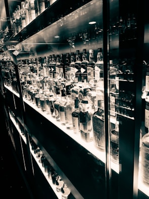 A well-lit shelf displaying an extensive collection of various liquor bottles organized neatly. The reflective surfaces create a mirrored effect, enhancing the visual richness. The black and white tone adds an elegant and timeless feel.