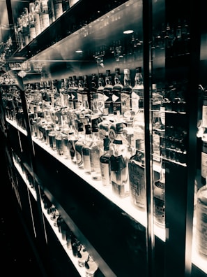 A well-lit shelf displaying an extensive collection of various liquor bottles organized neatly. The reflective surfaces create a mirrored effect, enhancing the visual richness. The black and white tone adds an elegant and timeless feel.
