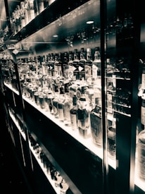 A well-lit shelf displaying an extensive collection of various liquor bottles organized neatly. The reflective surfaces create a mirrored effect, enhancing the visual richness. The black and white tone adds an elegant and timeless feel.