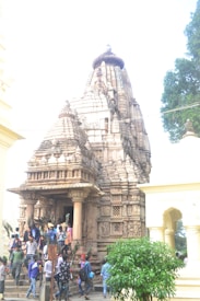 A group of people gathers at the entrance of an intricately carved stone temple. The temple structure features detailed sculptures and ornamental designs typical of traditional architecture. To the right, a small tree is visible, and the overall atmosphere suggests a cultural or religious gathering.