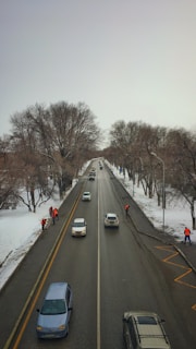 A snowy street scene captured from above shows a two-lane road with several vehicles driving in both directions. On the left side of the road, several workers wearing orange vests are performing maintenance tasks near the edge of the street. Leafless trees line both sides of the road, suggesting a winter setting.