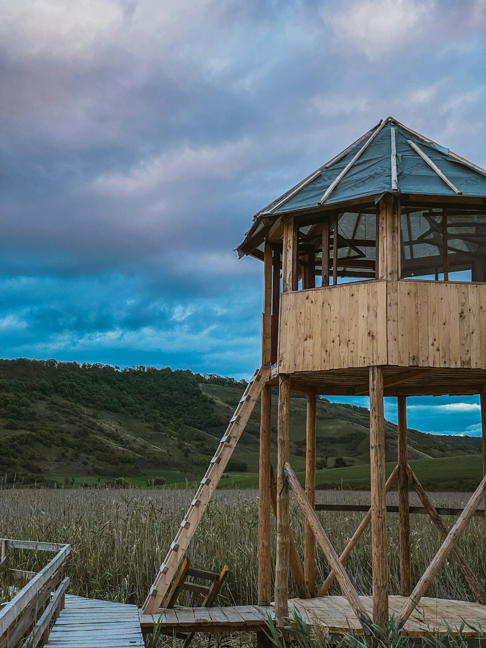 a wooden structure with a ladder on a wooden deck in front of a grassy hill