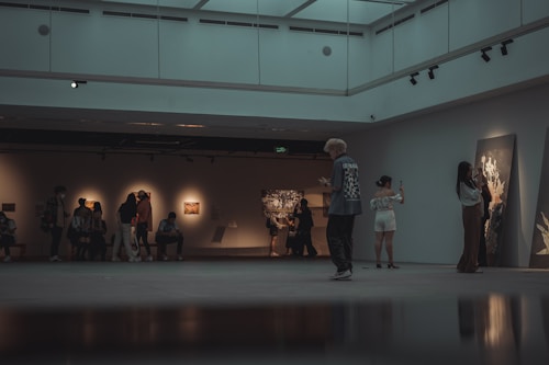 A spacious, dimly lit gallery features several groups of people observing and photographing artworks displayed on the walls. The mood is reflective as people engage with the exhibits, some seated on benches while others stand, capturing images on their phones.