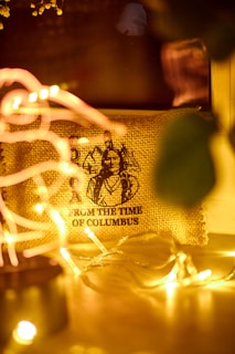 A rustic burlap canvas featuring a vintage illustration with the text 'FROM THE TIME OF COLUMBUS' partially visible. It is surrounded by soft, warm yellow fairy lights creating a cozy and nostalgic atmosphere.