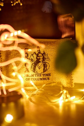 A rustic burlap canvas featuring a vintage illustration with the text 'FROM THE TIME OF COLUMBUS' partially visible. It is surrounded by soft, warm yellow fairy lights creating a cozy and nostalgic atmosphere.