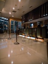 A modern hotel reception area featuring a sleek design. The counter is equipped with digital displays and several computers. The ambient lighting is warm, with pendant lights hanging from the ceiling. Decorative ropes guide visitors along the polished, reflective floor. There is a potted orchid on the counter, adding a touch of elegance.
