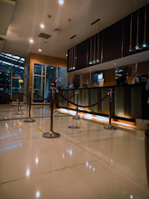 A modern hotel reception area featuring a sleek design. The counter is equipped with digital displays and several computers. The ambient lighting is warm, with pendant lights hanging from the ceiling. Decorative ropes guide visitors along the polished, reflective floor. There is a potted orchid on the counter, adding a touch of elegance.