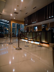 A modern hotel reception area featuring a sleek design. The counter is equipped with digital displays and several computers. The ambient lighting is warm, with pendant lights hanging from the ceiling. Decorative ropes guide visitors along the polished, reflective floor. There is a potted orchid on the counter, adding a touch of elegance.