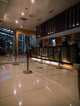 A modern hotel reception area featuring a sleek design. The counter is equipped with digital displays and several computers. The ambient lighting is warm, with pendant lights hanging from the ceiling. Decorative ropes guide visitors along the polished, reflective floor. There is a potted orchid on the counter, adding a touch of elegance.