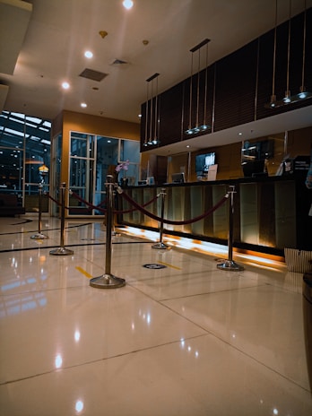 A modern hotel reception area featuring a sleek design. The counter is equipped with digital displays and several computers. The ambient lighting is warm, with pendant lights hanging from the ceiling. Decorative ropes guide visitors along the polished, reflective floor. There is a potted orchid on the counter, adding a touch of elegance.