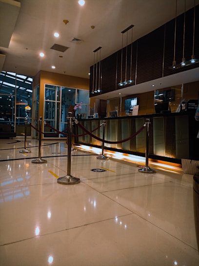 A modern hotel reception area featuring a sleek design. The counter is equipped with digital displays and several computers. The ambient lighting is warm, with pendant lights hanging from the ceiling. Decorative ropes guide visitors along the polished, reflective floor. There is a potted orchid on the counter, adding a touch of elegance.