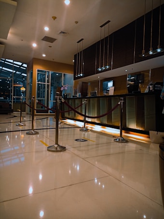 A modern hotel reception area featuring a sleek design. The counter is equipped with digital displays and several computers. The ambient lighting is warm, with pendant lights hanging from the ceiling. Decorative ropes guide visitors along the polished, reflective floor. There is a potted orchid on the counter, adding a touch of elegance.