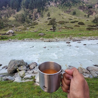 A metal cup filled with a warm beverage is held by a hand in the foreground, with a mountainous landscape in the background. The scene includes a flowing river with rocks on its banks, surrounded by lush greenery and distant hills covered with trees.