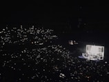 A large concert hall is filled with an audience holding up lights, such as smartphones, creating a sparkling effect throughout the darkened venue. A brightly lit stage features performers and illuminated square structures, providing the main source of light and focus.