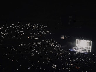 A large concert hall is filled with an audience holding up lights, such as smartphones, creating a sparkling effect throughout the darkened venue. A brightly lit stage features performers and illuminated square structures, providing the main source of light and focus.