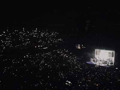 A large concert hall is filled with an audience holding up lights, such as smartphones, creating a sparkling effect throughout the darkened venue. A brightly lit stage features performers and illuminated square structures, providing the main source of light and focus.