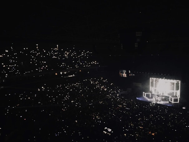 A large concert hall is filled with an audience holding up lights, such as smartphones, creating a sparkling effect throughout the darkened venue. A brightly lit stage features performers and illuminated square structures, providing the main source of light and focus.