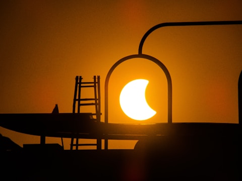 A solar eclipse is captured in silhouette surrounded by dark structures, possibly part of a building or equipment. The sun appears partially covered, creating a dramatic crescent shape against an orange skyline.