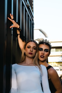 Two women are standing close to each other in front of a metallic background. One woman is wearing a white outfit with minimalistic makeup and red lipstick, while the other woman is dressed in black with dramatic eye makeup and a headpiece resembling a crown. They appear to be performing or posing for a thematic photo shoot.