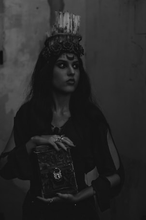 A woman with long dark hair wears a crown made of candles and floral decorations. Her makeup is dramatic with dark eye shadows, highlighting an enigmatic and intense expression. She is holding a textured, ornate book with a decorative clasp, and her fingers have multiple rings, enhancing a mystical or gothic appearance. The background is dimly lit, adding to the mysterious ambiance.
