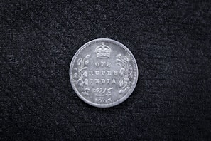 A vintage coin with the inscription 'ONE RUPEE INDIA 1905' and a crown symbol at the top. Decorative floral patterns adorn the sides. The coin rests on a textured black surface.