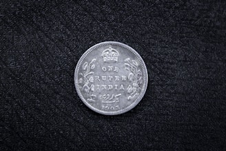 A vintage coin with the inscription 'ONE RUPEE INDIA 1905' and a crown symbol at the top. Decorative floral patterns adorn the sides. The coin rests on a textured black surface.