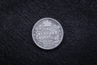 A vintage coin with the inscription 'ONE RUPEE INDIA 1905' and a crown symbol at the top. Decorative floral patterns adorn the sides. The coin rests on a textured black surface.