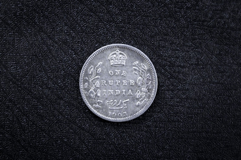 A vintage coin with the inscription 'ONE RUPEE INDIA 1905' and a crown symbol at the top. Decorative floral patterns adorn the sides. The coin rests on a textured black surface.