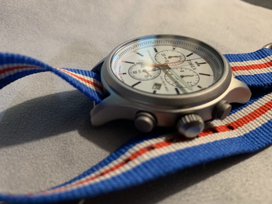 A close-up of a wristwatch with a round, silver case and a white dial featuring multiple sub-dials. The watch has a fabric strap in blue, white, and red stripes, creating a sporty and casual look. The surface under the watch appears soft and textured, possibly a piece of fabric or upholstery.
