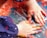 Two small hands covered in vibrant paint are actively engaged in finger painting on a surface, creating a swirl of colors. The background features a mixture of orange, blue, and other hues, suggesting a playful and creative activity.