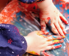 Two small hands covered in vibrant paint are actively engaged in finger painting on a surface, creating a swirl of colors. The background features a mixture of orange, blue, and other hues, suggesting a playful and creative activity.