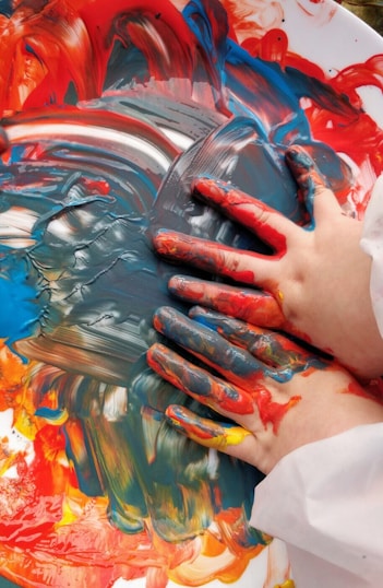 Two hands covered in vibrant paint smudges swirl a mix of red, blue, yellow, and orange colors on a surface, creating a vivid and dynamic abstract pattern.