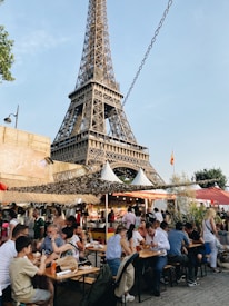 A busy outdoor caf&eacute; scene at the base of the Eiffel Tower, where people are seated around picnic-style wooden tables enjoying food and drinks. Behind them, multiple food and drink stalls can be seen, each with colorful signage. The atmosphere is lively and bustling, with people of various ages engaged in conversation. The iconic Eiffel Tower looms prominently in the background, standing tall against a clear blue sky.