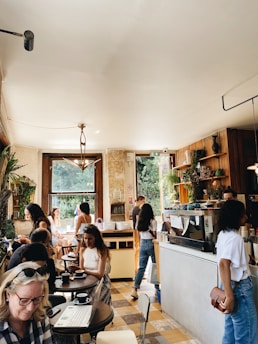 A bustling café with several people engaged in various activities. Some are sitting at tables working on laptops or conversing, while others are standing near the counter. The space is sunny and features wooden accents, plants, and a large window offering a view of greenery outside.