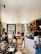A bustling café with several people engaged in various activities. Some are sitting at tables working on laptops or conversing, while others are standing near the counter. The space is sunny and features wooden accents, plants, and a large window offering a view of greenery outside.