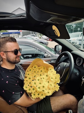 A person with sunglasses is sitting in the driver's seat of a car, holding a large bouquet of yellow flowers. The interior of the car is visible, including a BMW logo on the steering wheel. Several cars are parked outside in the background.