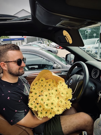 A person with sunglasses is sitting in the driver's seat of a car, holding a large bouquet of yellow flowers. The interior of the car is visible, including a BMW logo on the steering wheel. Several cars are parked outside in the background.