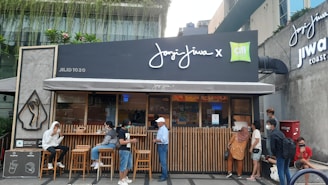A small coffee shop with a modern design and outdoor seating. Several people are gathered outside, some wearing face masks, indicating health precautions. The storefront features a dark facade with large signage, and lush greenery is visible above.