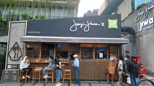 A small coffee shop with a modern design and outdoor seating. Several people are gathered outside, some wearing face masks, indicating health precautions. The storefront features a dark facade with large signage, and lush greenery is visible above.
