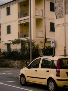 A small yellow car is parked on a street next to a yellow, three-story residential building with balconies and closed shutters. A stone wall partially covers the right side of the building. There is a street sign and a pole nearby, along with some greenery behind a fence.